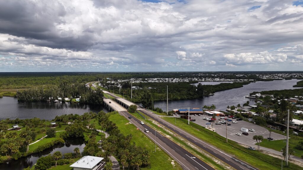 Aerial view of a roadway, bridge, waterway, and surrounding natural landscape beneath cloudy skies
