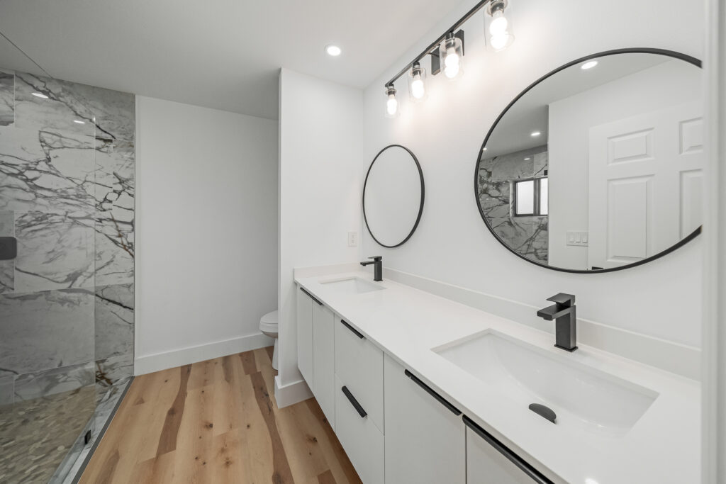 Bright modern bathroom with a double vanity, round mirrors, walk-in shower, and wood-look flooring
